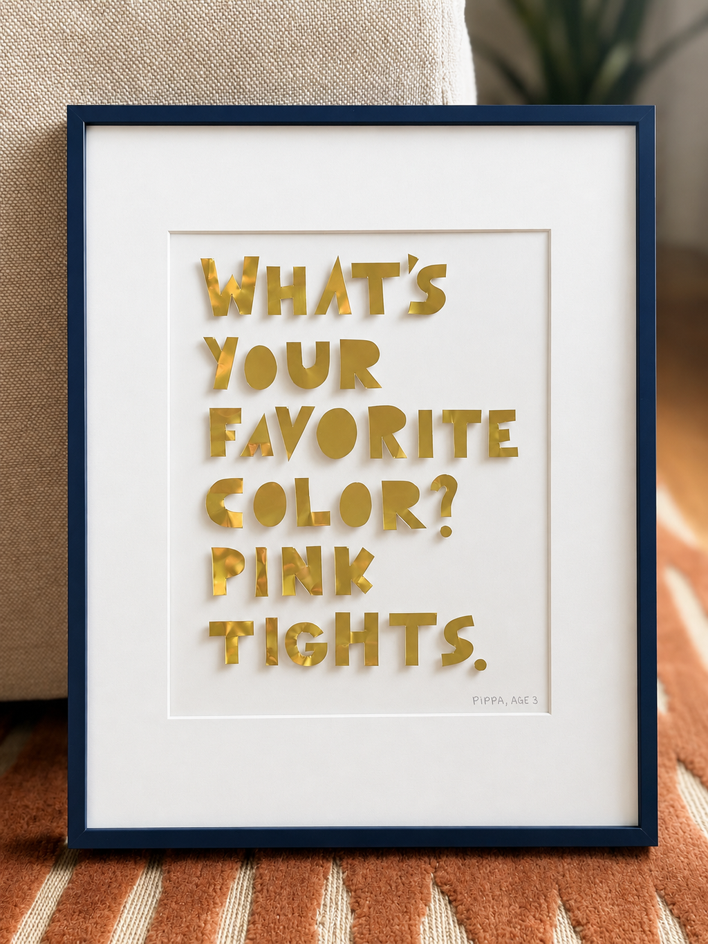 A quote piece in a navy frame in front of a sofa, gold hand-cut metal letters reading what's your favorite color, pink tights.