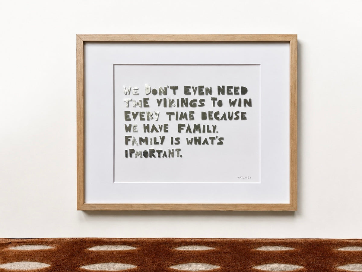 A framed quote piece in a natural oak frame, hand-cut metal letters reading a quote about family on cream cardstock, hung on a white wall.