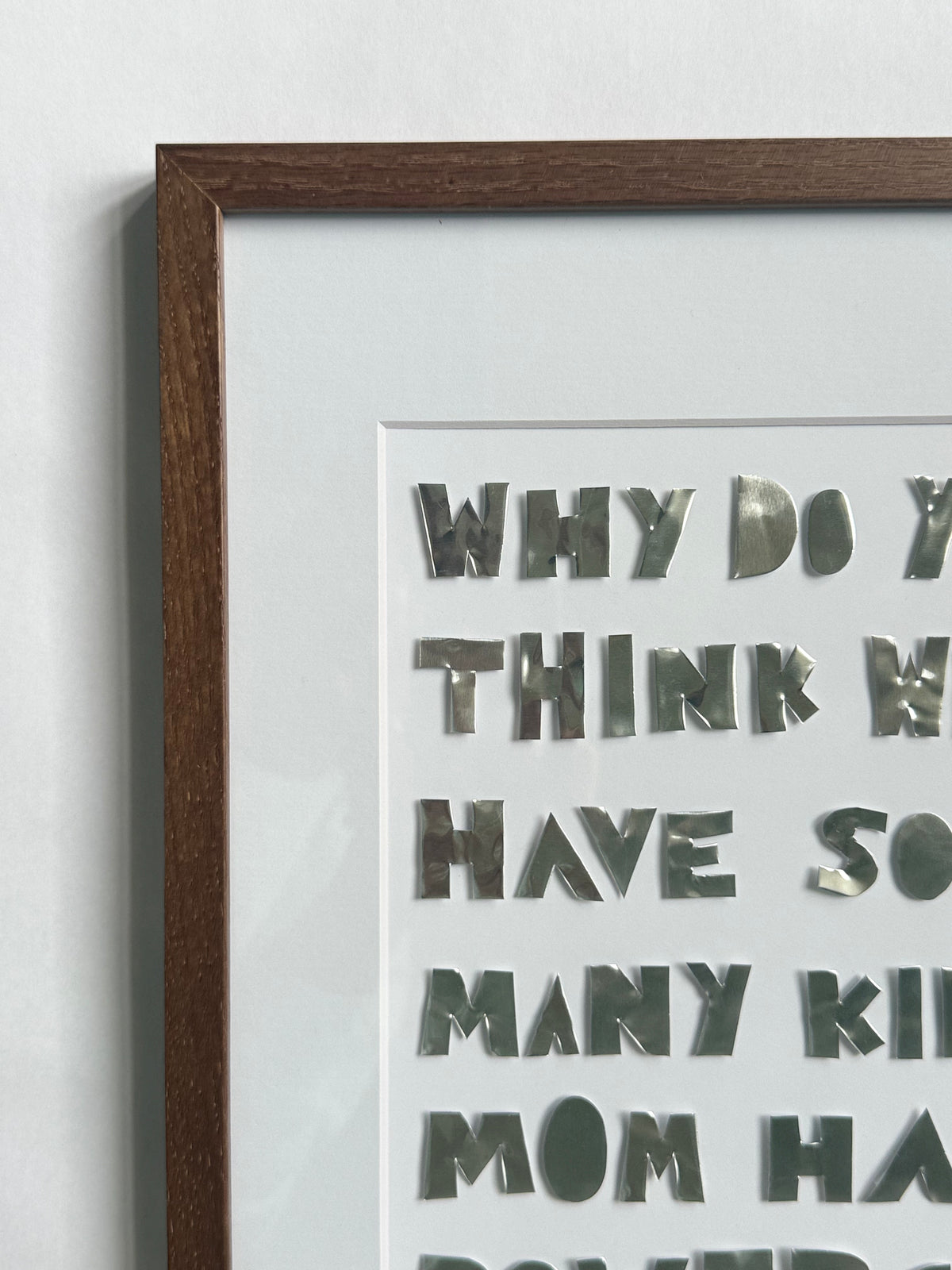 Close-up of a quote piece in a dark wood frame, silver hand-cut metal letters on cream cardstock reading why do you think we have so many kids, mom has powers.