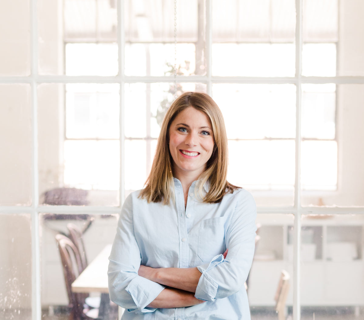 Lucy, founder of Pippy + Funk, smiling with her arms crossed in a light blue shirt in front of a bright window.