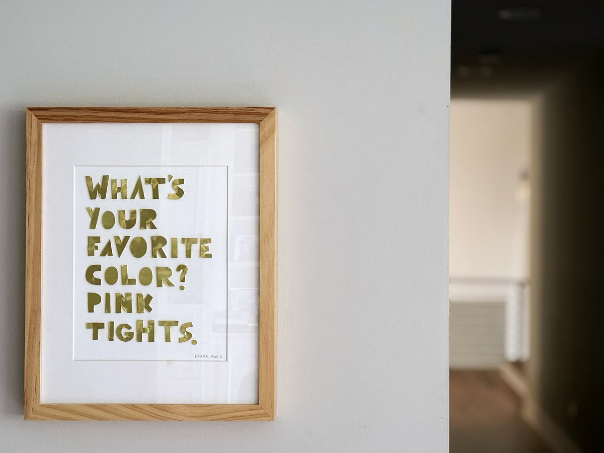 A framed quote piece in a natural oak frame, gold hand-cut metal letters reading what's your favorite color, pink tights, hung on a wall by a doorway.