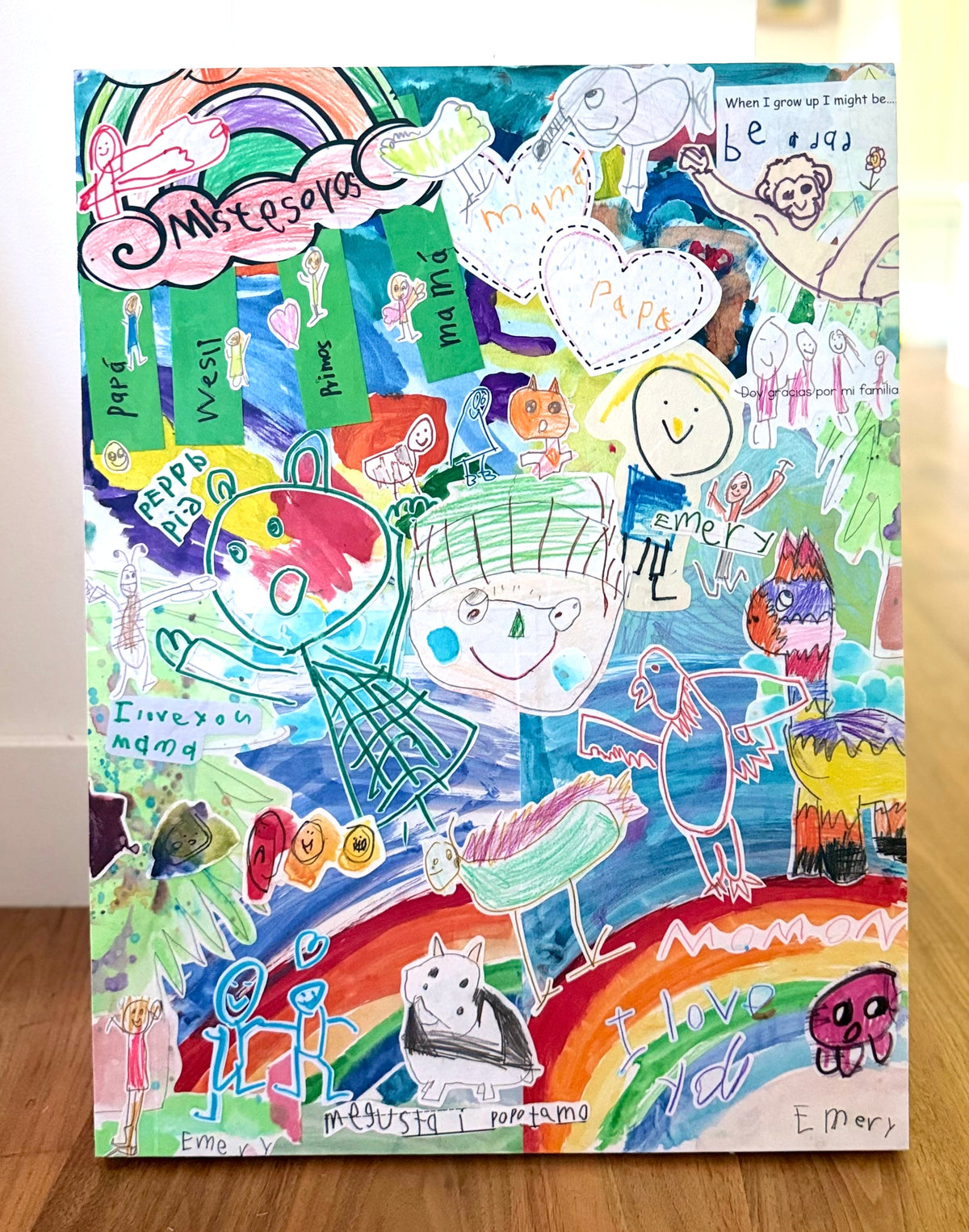 A finished custom collage of a child's artwork, with rainbows, characters, and handwriting layered on a basswood panel, standing on a wood floor.